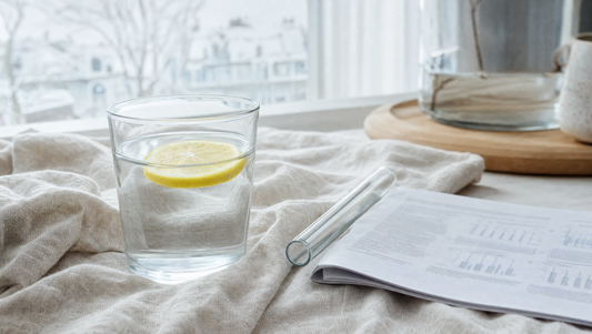 Glas water met citroenschijf naast laboratoriumglas en wetenschappelijk papier — detox-januari en leverontgifting onder de loep