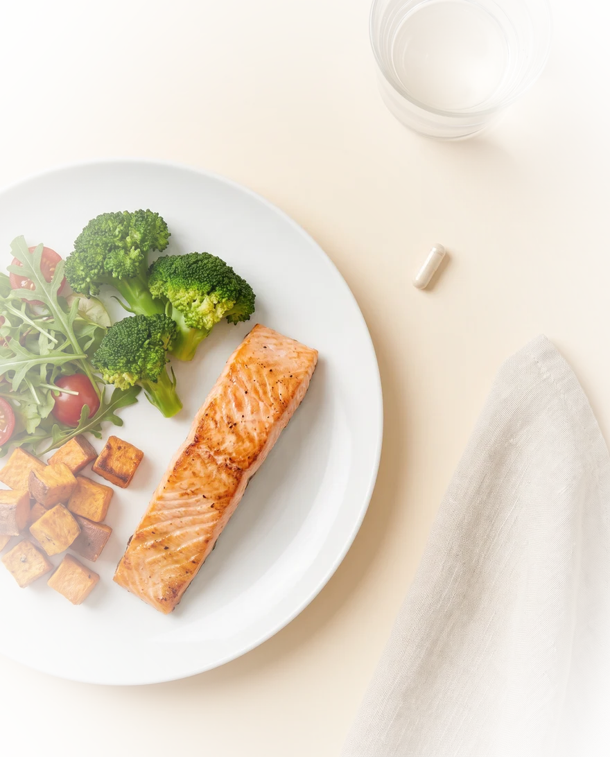 Eén capsule naast een bord met zalm, broccoli en zoete aardappel, van bovenaf
